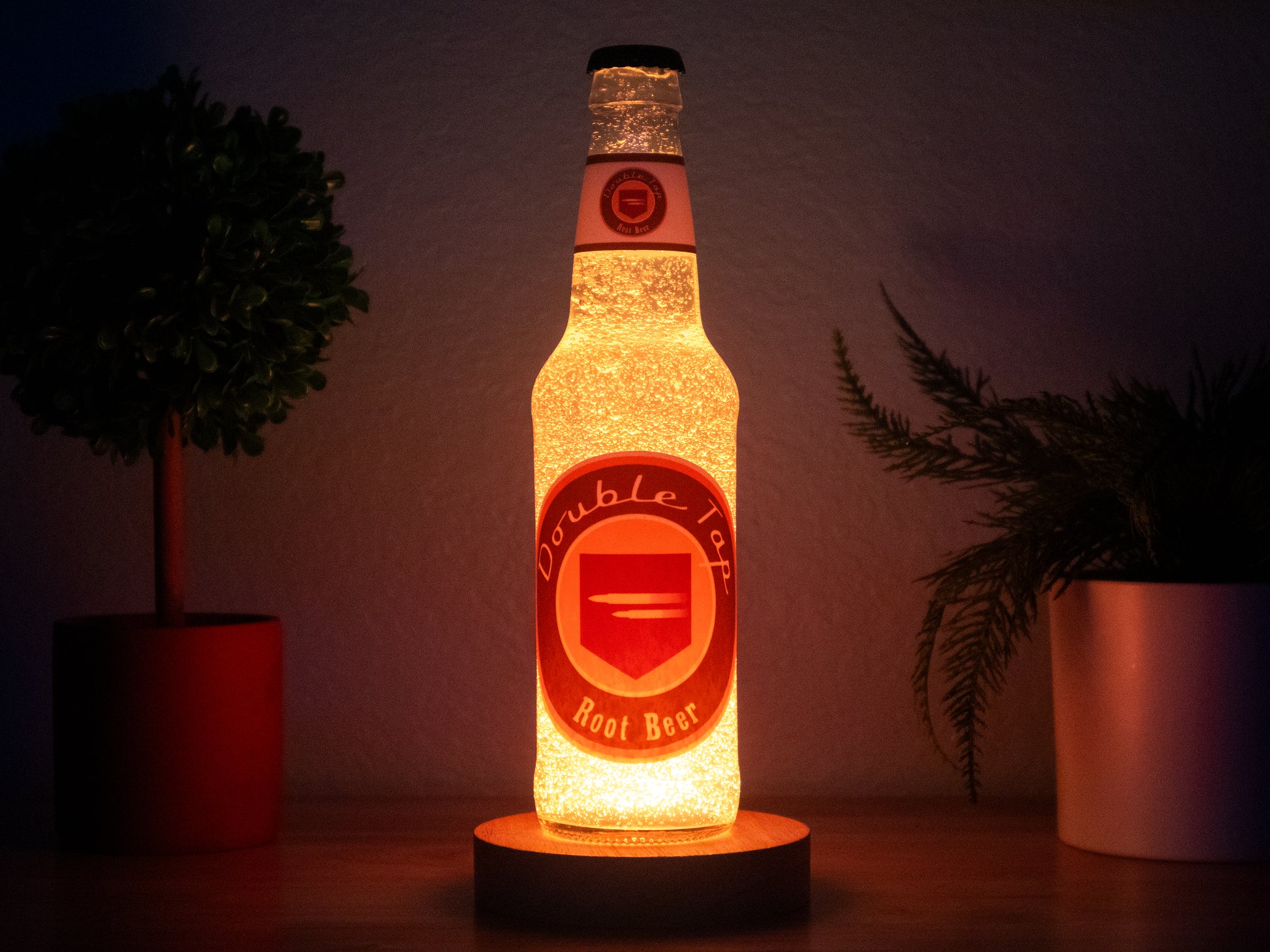 A Double Tap Root Beer Perk a Cola Bottle Light by Bottle Light Company sits on wood beside two potted plants in dim light—the perfect tribute for Call of Duty Zombies fans.
