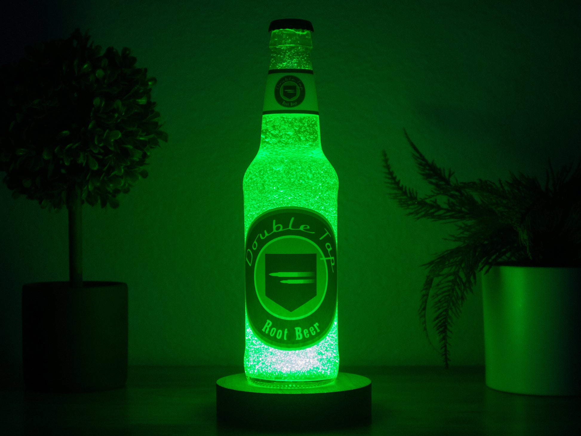 The Bottle Light Company’s Double Tap Root Beer Perk a Cola Bottle Light, inspired by Call of Duty Zombies, emits a vibrant green glow from below. It’s pictured on a dark surface between two small potted plants with a moody backdrop.