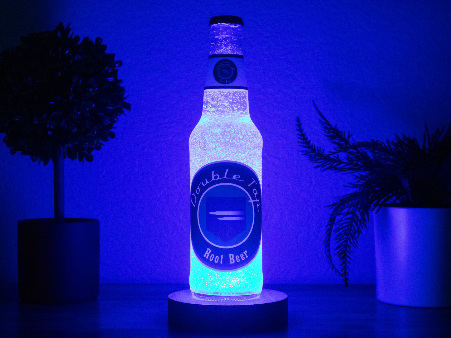 The Double Tap Root Beer Perk a Cola Bottle Light by Bottle Light Company glows blue on a table between two potted plants, evoking the iconic Call of Duty Zombies drink against a dim background.