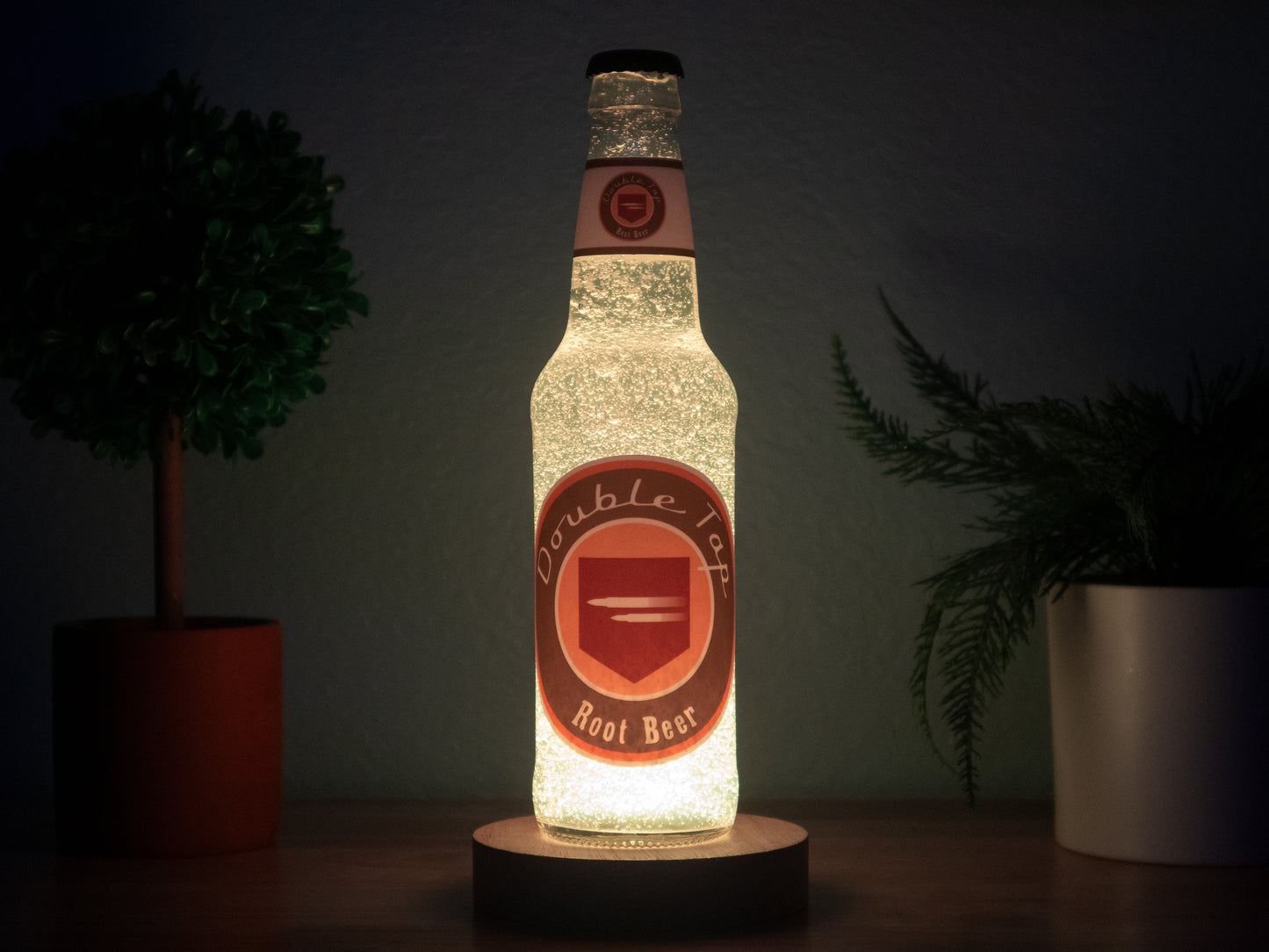 The Bottle Light Company’s Double Tap Root Beer Perk a Cola Bottle Light glows on a wooden surface between two small potted plants, evoking Call of Duty Zombies against a dark backdrop.