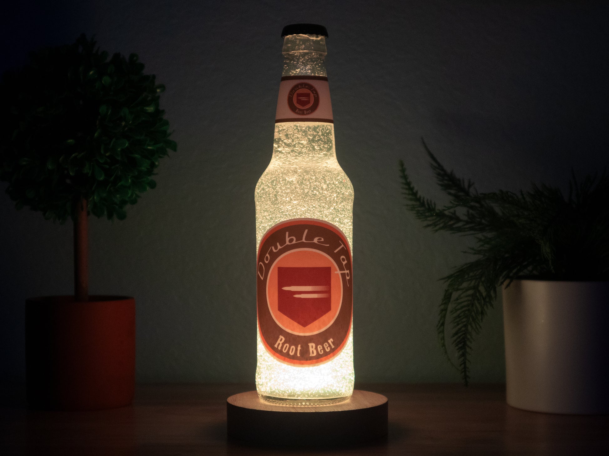 The Bottle Light Company’s Double Tap Root Beer Perk a Cola Bottle Light glows on a wooden surface between two small potted plants, evoking Call of Duty Zombies against a dark backdrop.