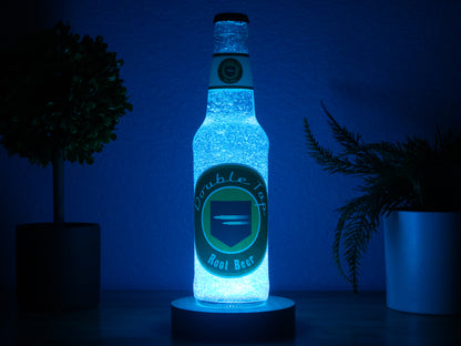 A glowing blue Double Tap Root Beer Perk a Cola Bottle Light by Bottle Light Company stands on a table between two potted plants, its illumination creating a dramatic Call of Duty Zombies-inspired effect against a dark blue background.