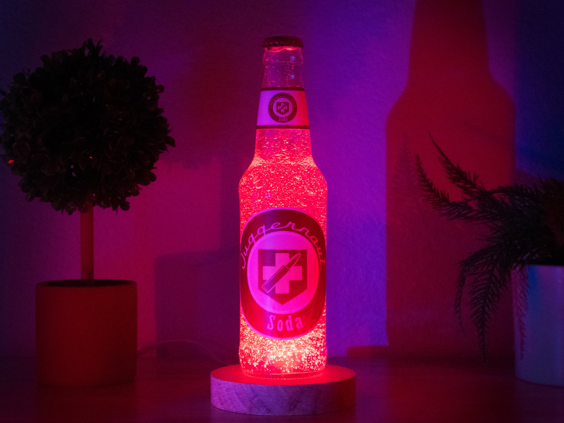The Juggernog Perk a Cola Bottle Light by Bottle Light Company, inspired by Call of Duty Zombies, glows red on a circular base. It’s displayed on wood with two potted plants in the background.