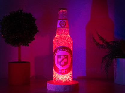 The Juggernog Perk a Cola Bottle Light by Bottle Light Company, inspired by Call of Duty Zombies, glows red on a circular base. It’s displayed on wood with two potted plants in the background.