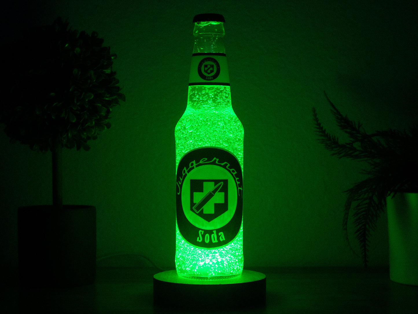 The Juggernog Perk a Cola Bottle Light by Bottle Light Company glows bright green, inspired by Call of Duty Zombies, and sits on a dark surface with blurred potted plants in the background.