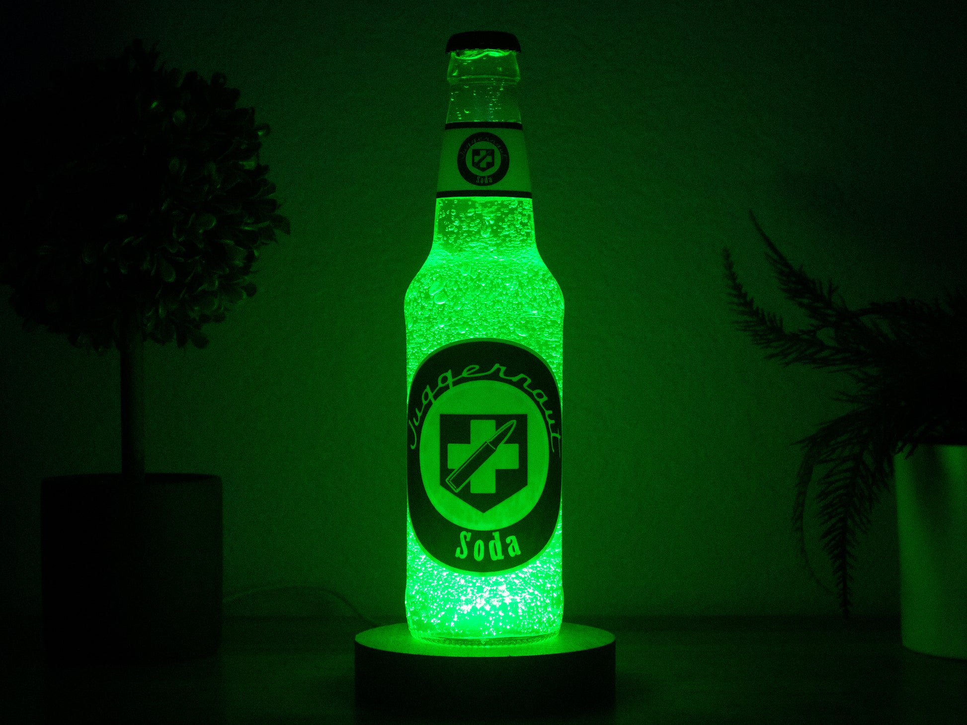 The Juggernog Perk a Cola Bottle Light by Bottle Light Company glows bright green, inspired by Call of Duty Zombies, and sits on a dark surface with blurred potted plants in the background.