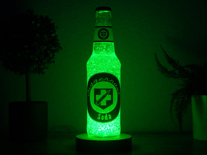 The Juggernog Perk a Cola Bottle Light by Bottle Light Company glows bright green, inspired by Call of Duty Zombies, and sits on a dark surface with blurred potted plants in the background.