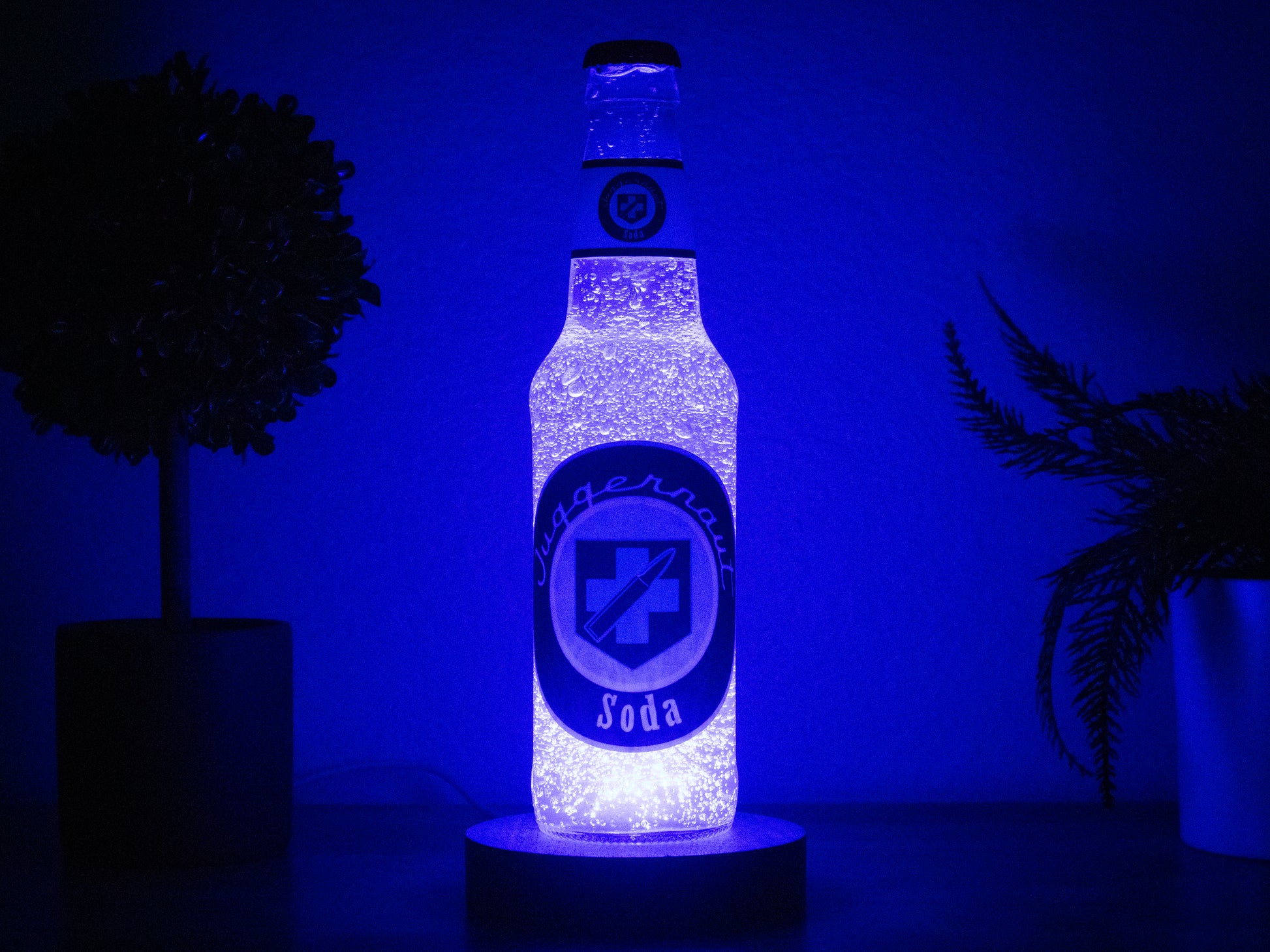The Juggernog Perk a Cola Bottle Light by Bottle Light Company glows on a round base between two potted plants in a dimly lit room, inspired by Call of Duty Zombies and illuminated with an LED light.