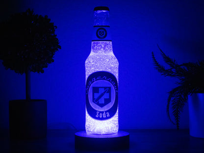 The Juggernog Perk a Cola Bottle Light by Bottle Light Company glows on a round base between two potted plants in a dimly lit room, inspired by Call of Duty Zombies and illuminated with an LED light.