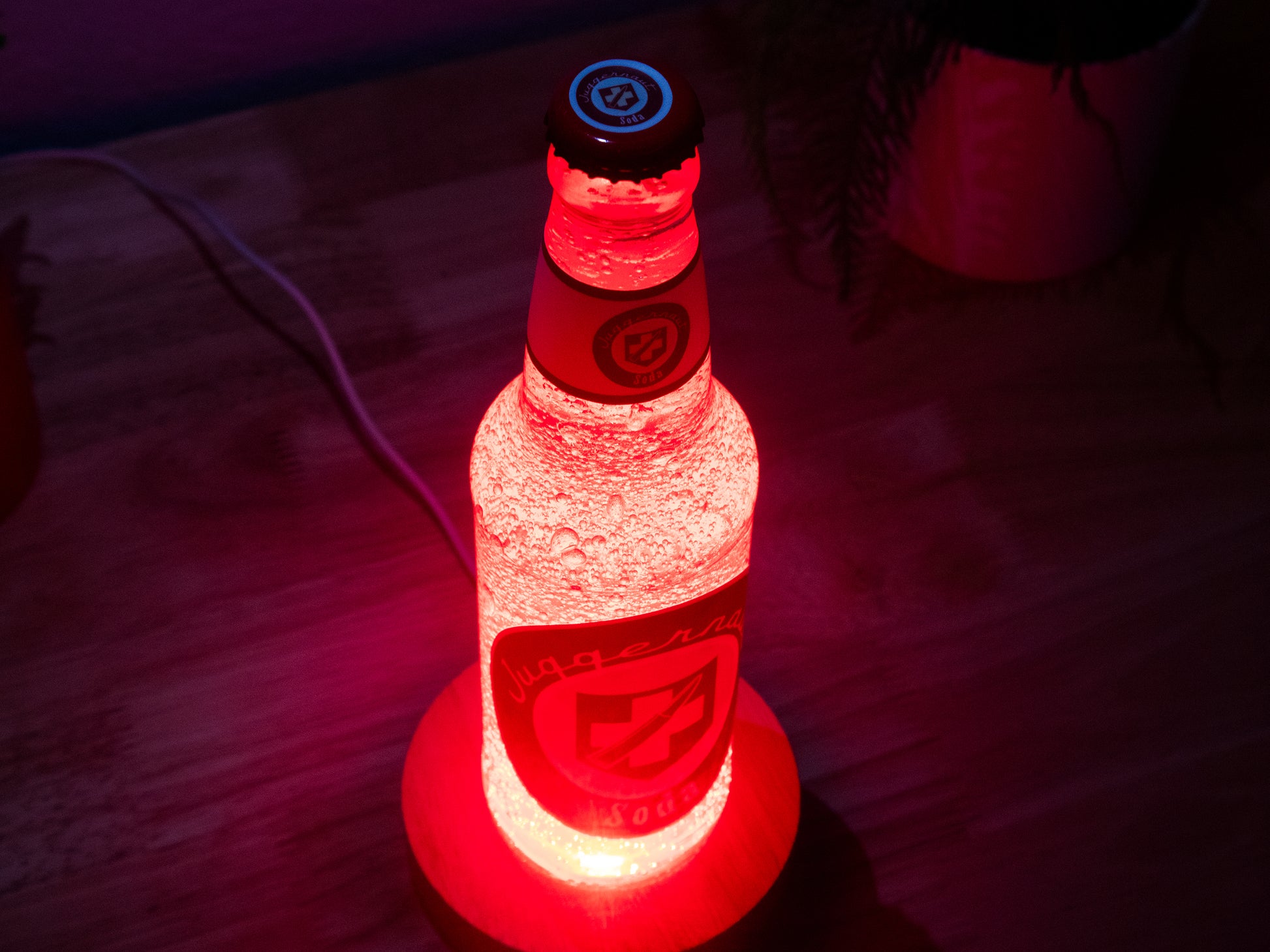 A Juggernog Perk a Cola Bottle Light by Bottle Light Company glows red on a wooden surface, evoking the Call of Duty Zombies item, with a glimpse of a plant pot in the background.