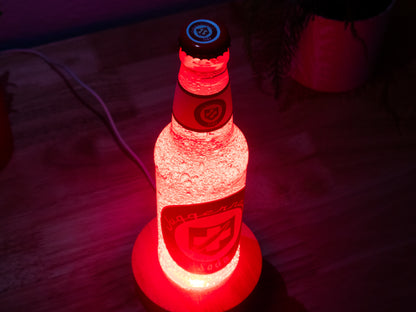 A Juggernog Perk a Cola Bottle Light by Bottle Light Company glows red on a wooden surface, evoking the Call of Duty Zombies item, with a glimpse of a plant pot in the background.