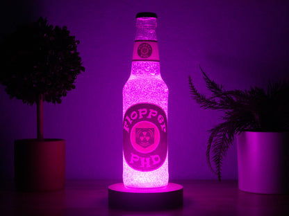The Bottle Light Company’s PHD Flopper Perk a Cola Bottle Light glows bright pink on a wooden surface between two potted plants against a purple background—a must-have collectible for Call of Duty Zombies fans.