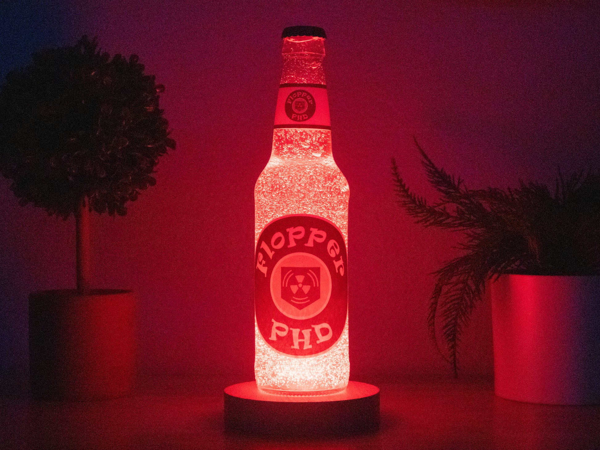 The Bottle Light Company’s PHD Flopper Perk a Cola Bottle Light glows red on a round base between two potted plants, making it a unique collectible decor piece inspired by Call of Duty Zombies.