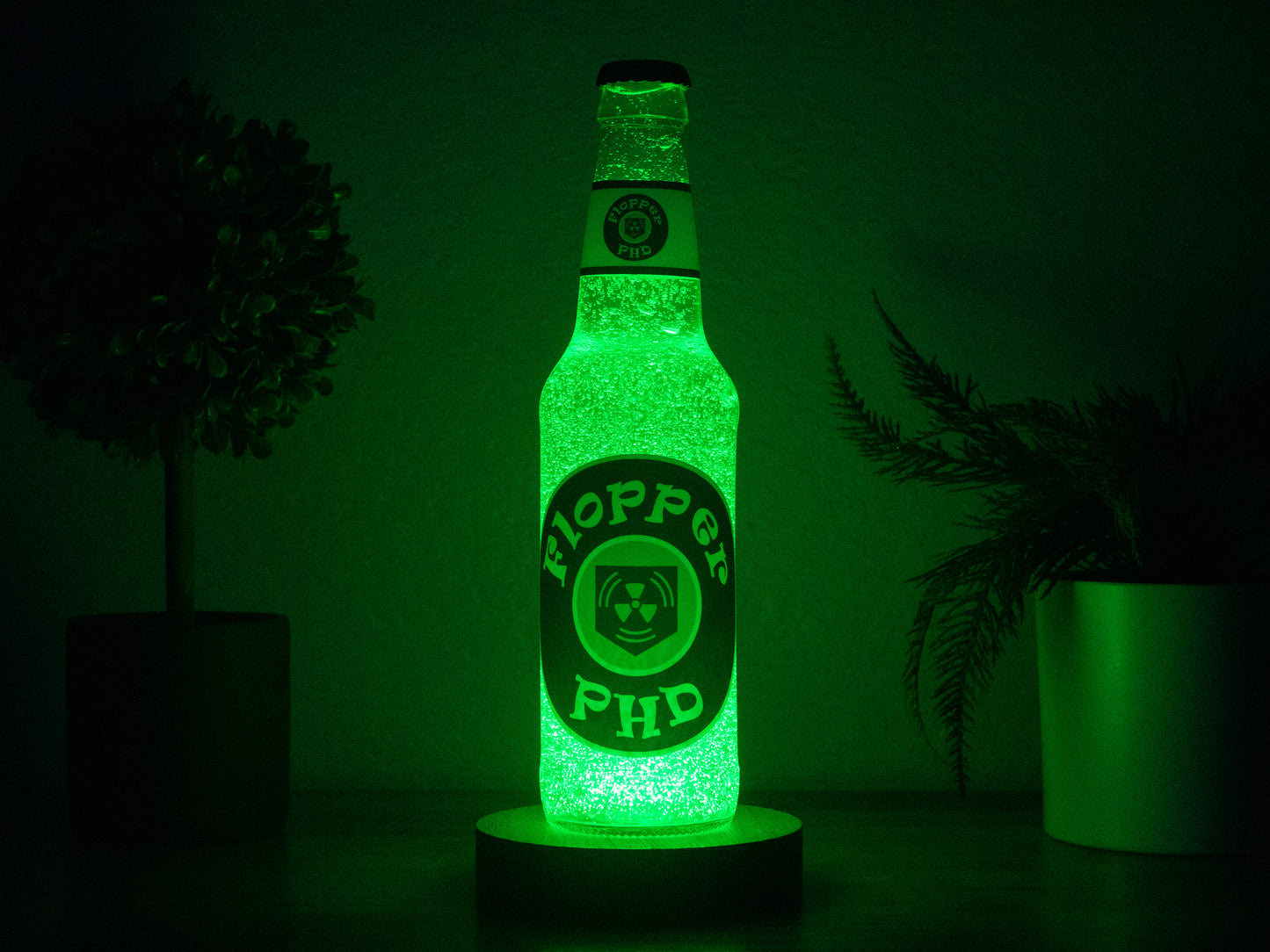 The PHD Flopper Perk a Cola Bottle Light by Bottle Light Company glows green between two plants, adding collectible Call of Duty Zombies charm to any dark room.
