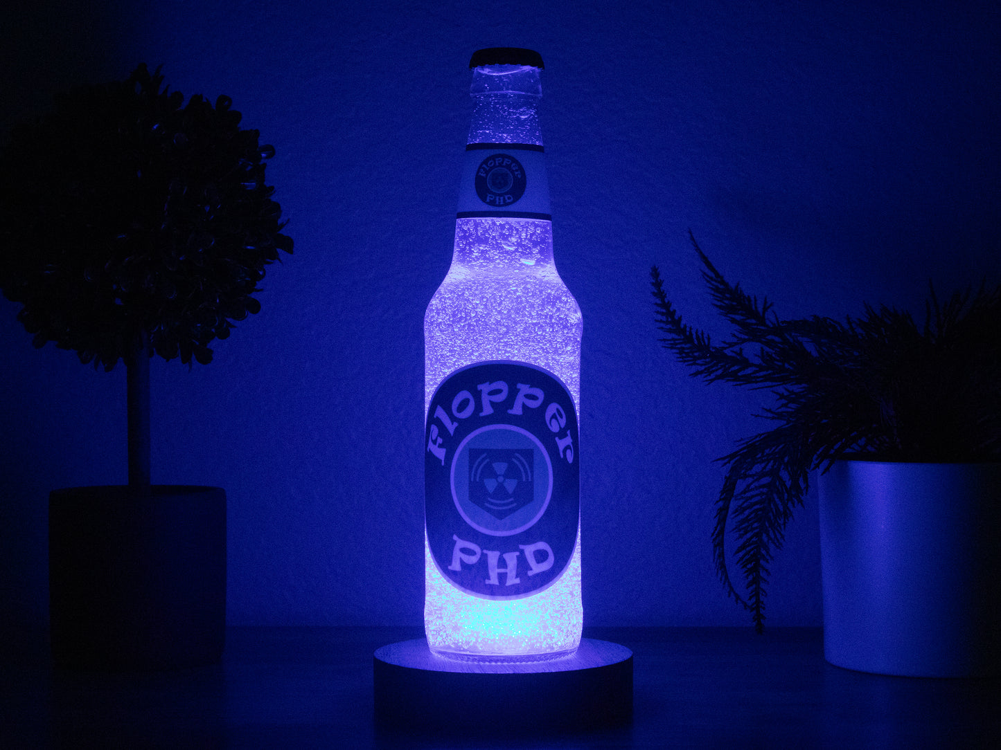 The PHD Flopper Perk a Cola Bottle Light by Bottle Light Company glows blue between two potted plants on a dark surface, inspired by Call of Duty Zombies and set against a deep blue background—perfect collectible decor.
