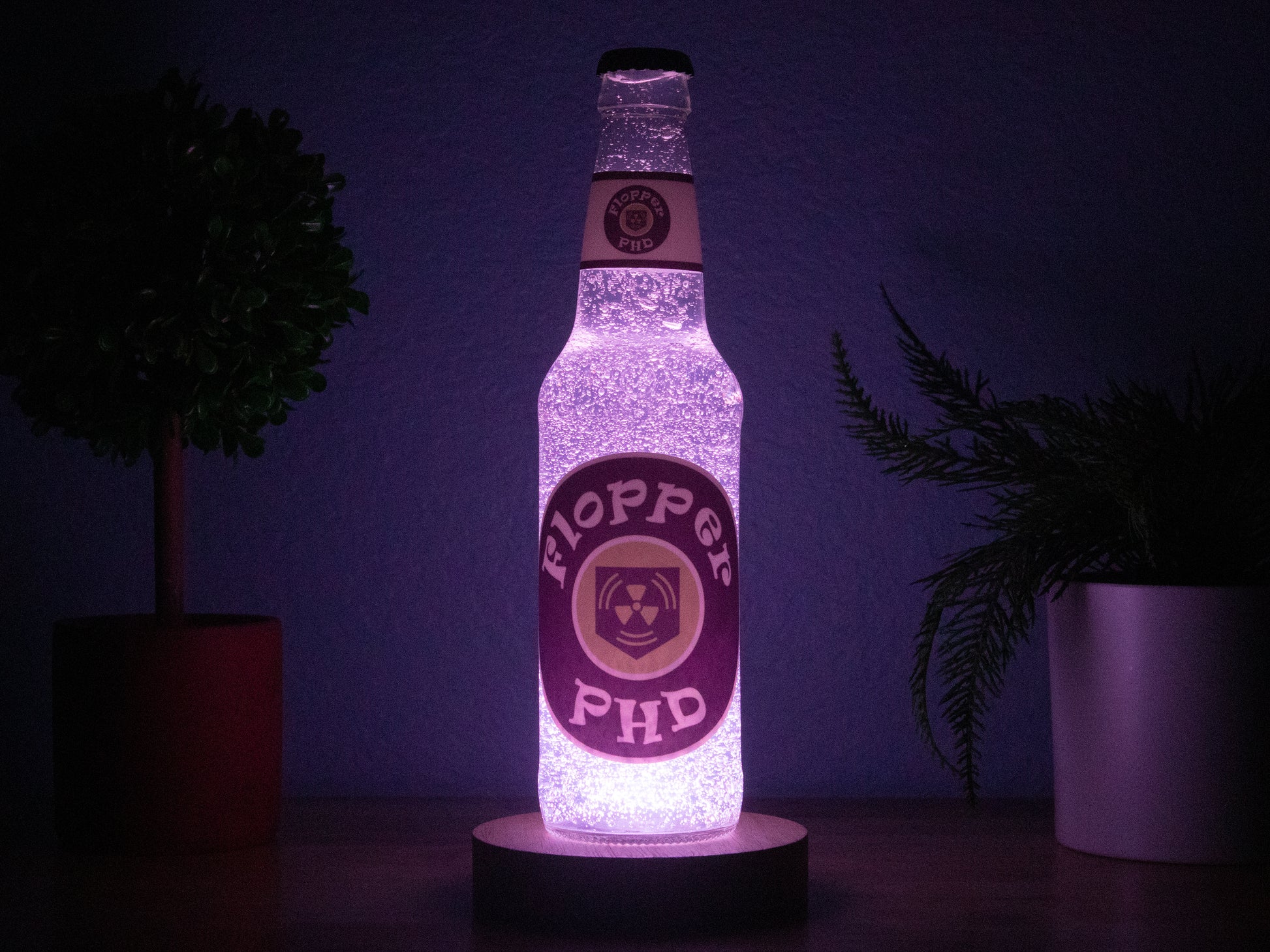 The PHD Flopper Perk a Cola Bottle Light by Bottle Light Company glows softly purple between potted plants—an essential collectible decor piece for any Call of Duty Zombies fan.