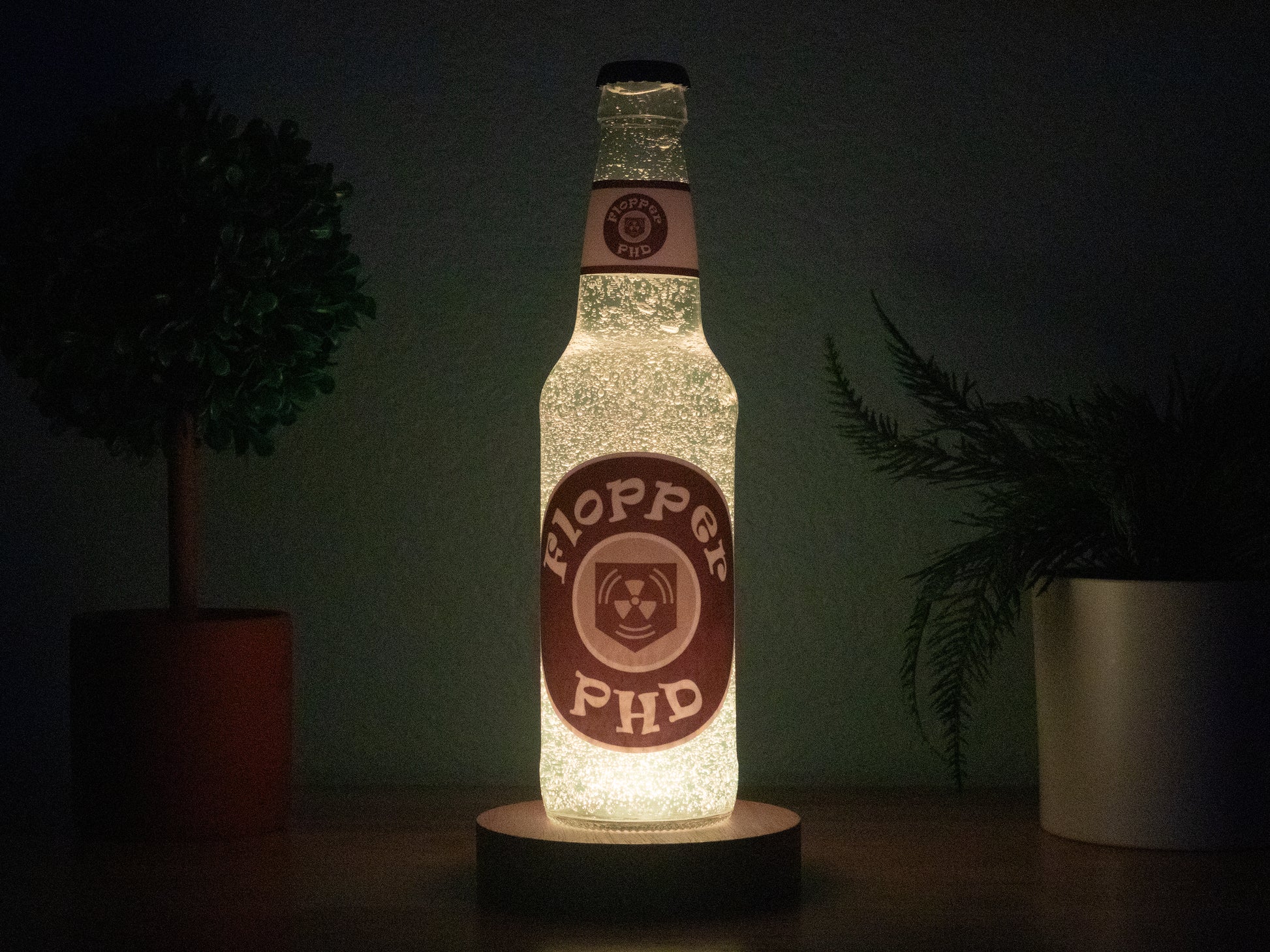The PHD Flopper Perk a Cola Bottle Light by Bottle Light Company glows white on a wooden surface between two potted plants—a perfect Call of Duty Zombies collectible for fans of the iconic perk.
