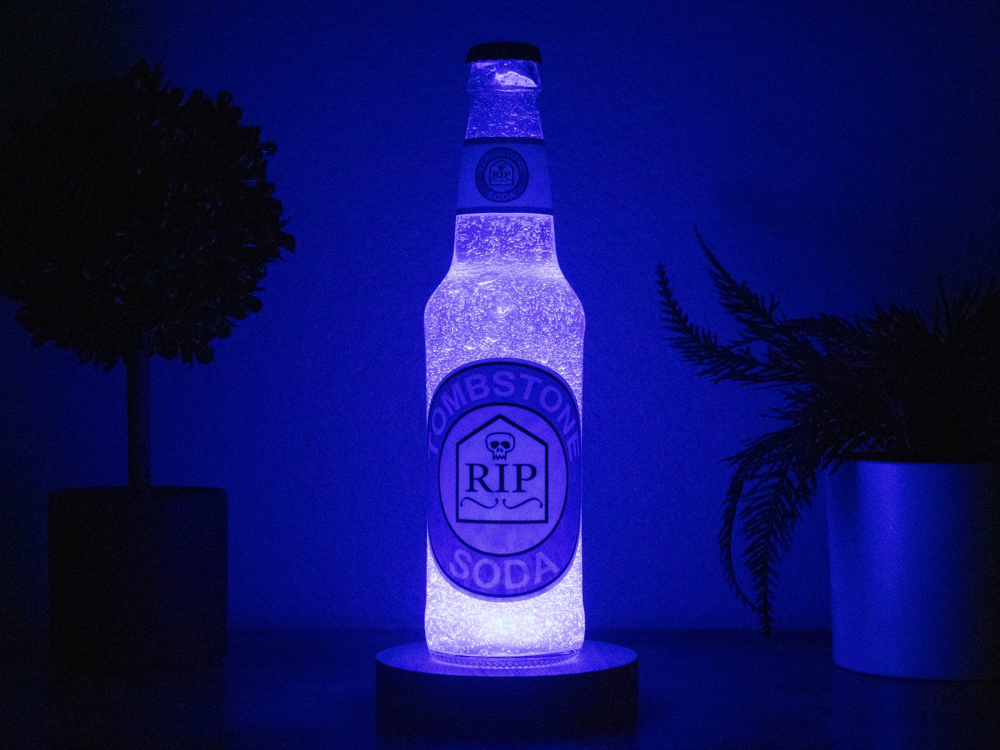 The Bottle Light Company’s Tombstone Perk a Cola Bottle Light, glowing with its RIP gravestone logo, is displayed between two potted plants on a dark surface—a striking collectible for Call of Duty Zombies fans.