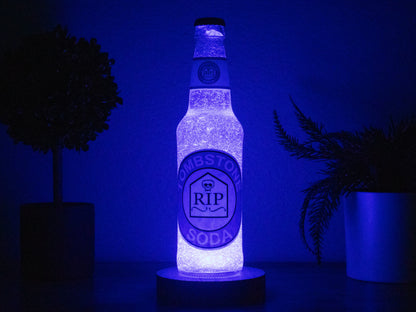 The Bottle Light Company’s Tombstone Perk a Cola Bottle Light, glowing with its RIP gravestone logo, is displayed between two potted plants on a dark surface—a striking collectible for Call of Duty Zombies fans.