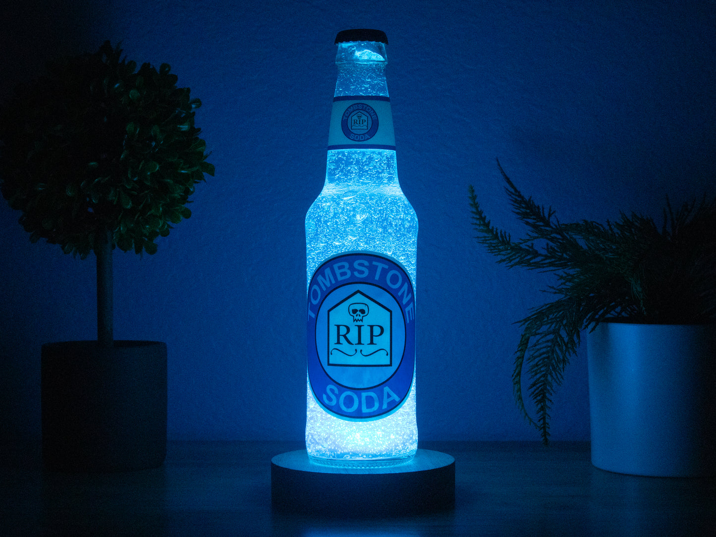 The Tombstone Perk a Cola Bottle Light by Bottle Light Company glows blue between two potted plants, casting cool illumination against a dark background. Inspired by Call of Duty Zombies Tombstone Soda perk.