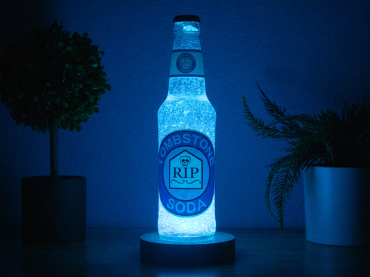 The Tombstone Perk a Cola Bottle Light by Bottle Light Company glows blue between two potted plants, casting cool illumination against a dark background. Inspired by Call of Duty Zombies Tombstone Soda perk.