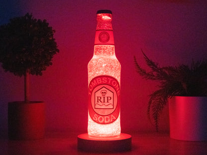 The Tombstone Perk a Cola Bottle Light by Bottle Light Company, featuring a glowing red RIP label with a skull, sits between two potted plants in dim light—a perfect Call of Duty Zombies collectible.