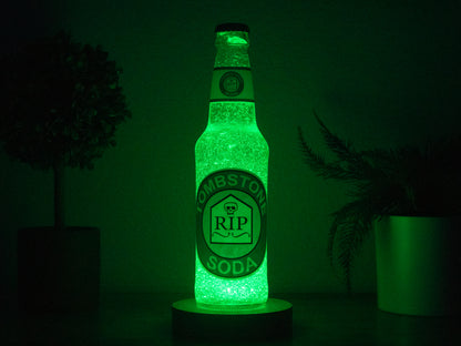 The Bottle Light Company’s Tombstone Perk a Cola Bottle Light glows green with a tombstone and RIP logo, standing between two potted plants in the dark.