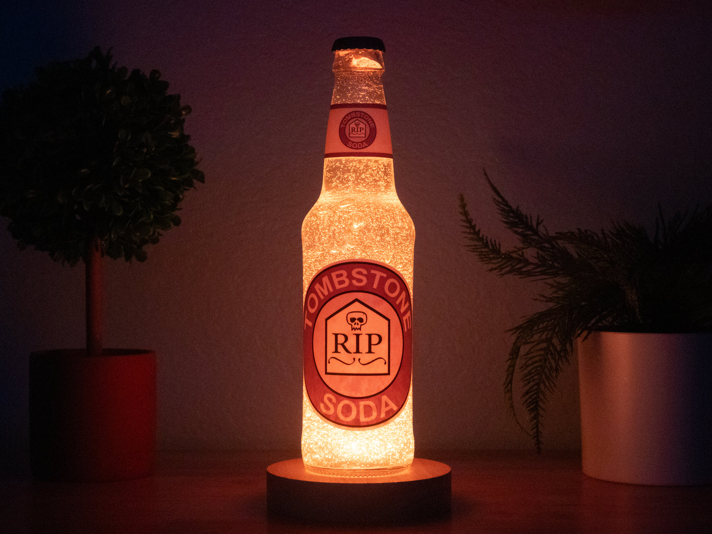 The Bottle Light Company Tombstone Perk a Cola Bottle Light glows orange on a wooden surface between two potted plants. Its RIP tombstone label makes it an ideal collectible for Call of Duty Zombies fans.