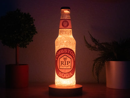 The Bottle Light Company Tombstone Perk a Cola Bottle Light glows orange on a wooden surface between two potted plants. Its RIP tombstone label makes it an ideal collectible for Call of Duty Zombies fans.