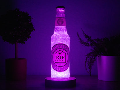 The Tombstone Perk a Cola Bottle Light by Bottle Light Company glows purple in the dark on a round base between potted plants—a unique Call of Duty Zombies collectible and LED bottle light.