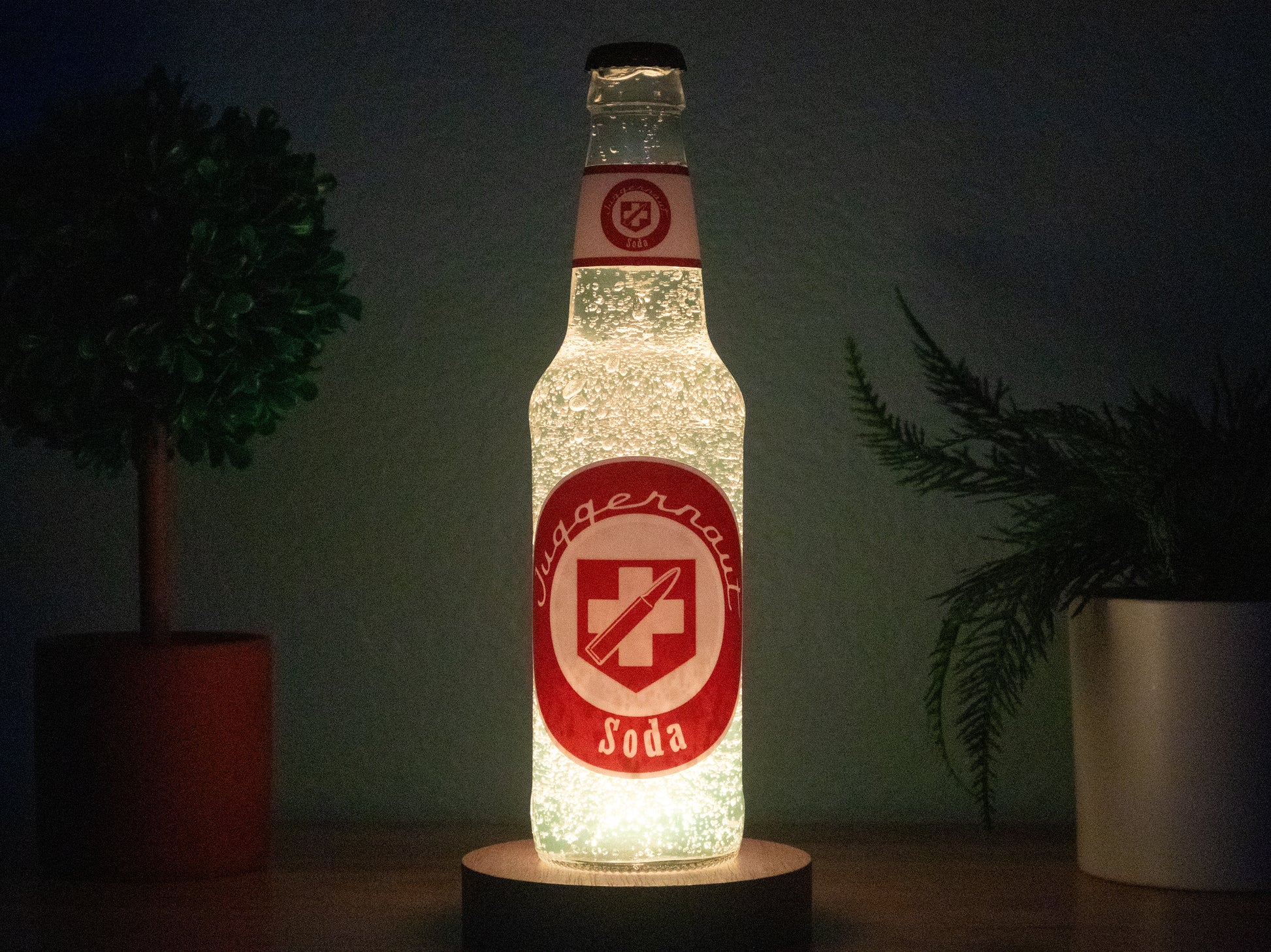 A Juggernog Perk a Cola Bottle Light by Bottle Light Company, glowing red and white, sits between two potted plants on a dark surface, illuminated from below and evoking the classic Call of Duty Zombies Perk-a-Cola.