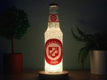 A Juggernog Perk a Cola Bottle Light by Bottle Light Company, glowing red and white, sits between two potted plants on a dark surface, illuminated from below and evoking the classic Call of Duty Zombies Perk-a-Cola.