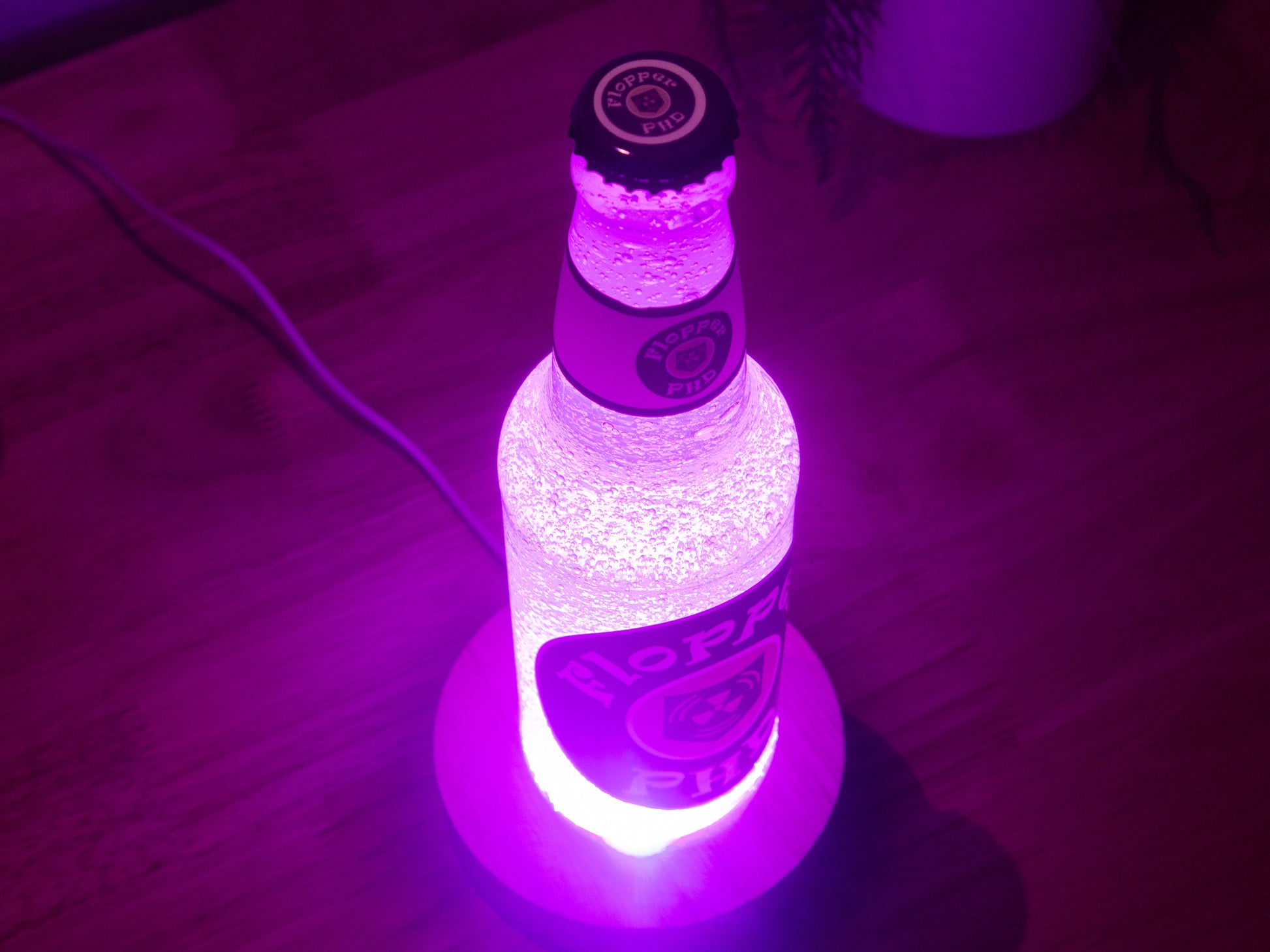 The Bottle Light Company’s PHD Flopper Perk a Cola Bottle Light glows with bright purple illumination on wood, creating collectible gamer décor reminiscent of Call of Duty Zombies. A cable and plant pot are also visible in the scene.